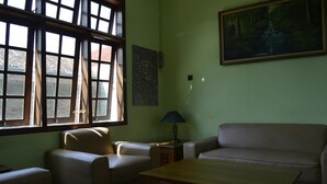 A room with a green wall, a painting, a window with blinds, a sofa, a chair, and a table. - Sartika Hotel (Yogyakarta)