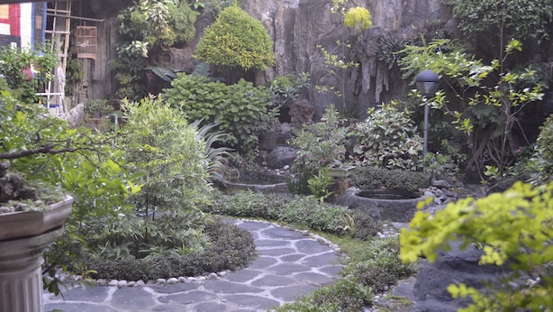 A garden with a stone pathway, various plants, and a rustic building with a red roof. - Sartika Hotel (Yogyakarta)