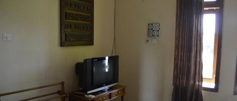 A room with a television on a wooden stand, a patterned wall hanging, and a window with curtains.