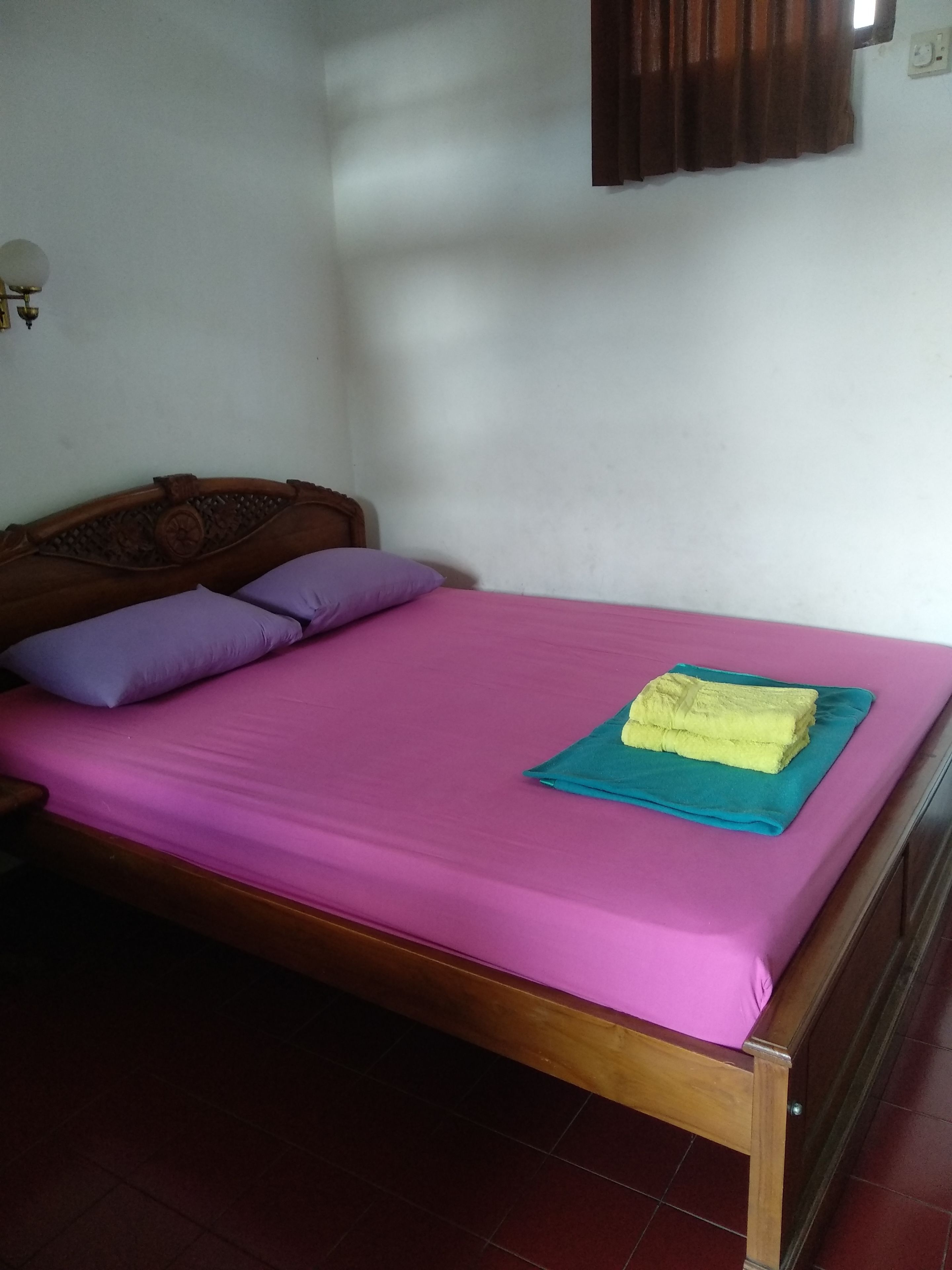 A single bed with a wooden headboard, a pink bedspread, and two purple pillows.