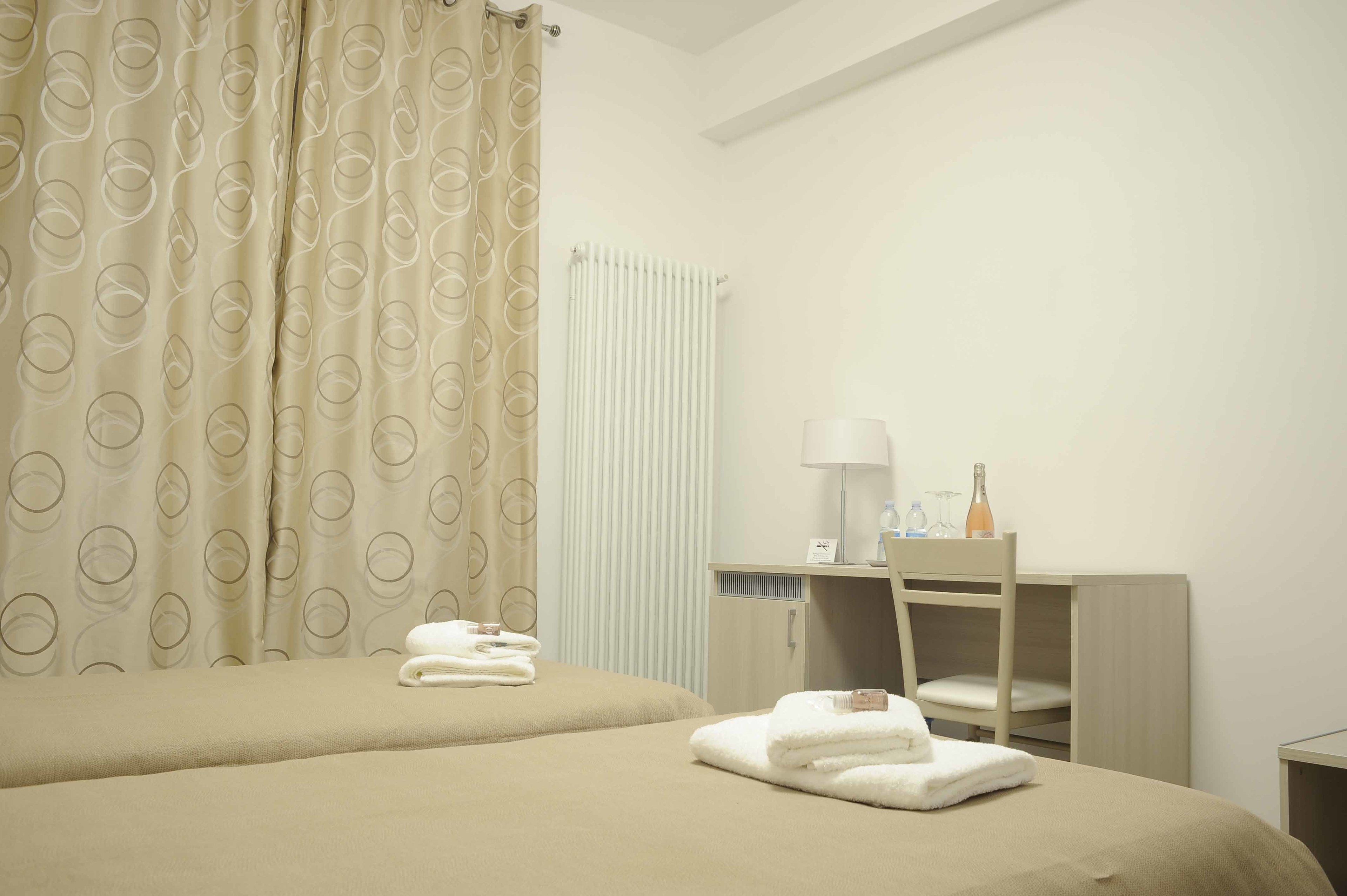 A hotel room with two beds, a desk, a chair, a radiator, and a lamp.