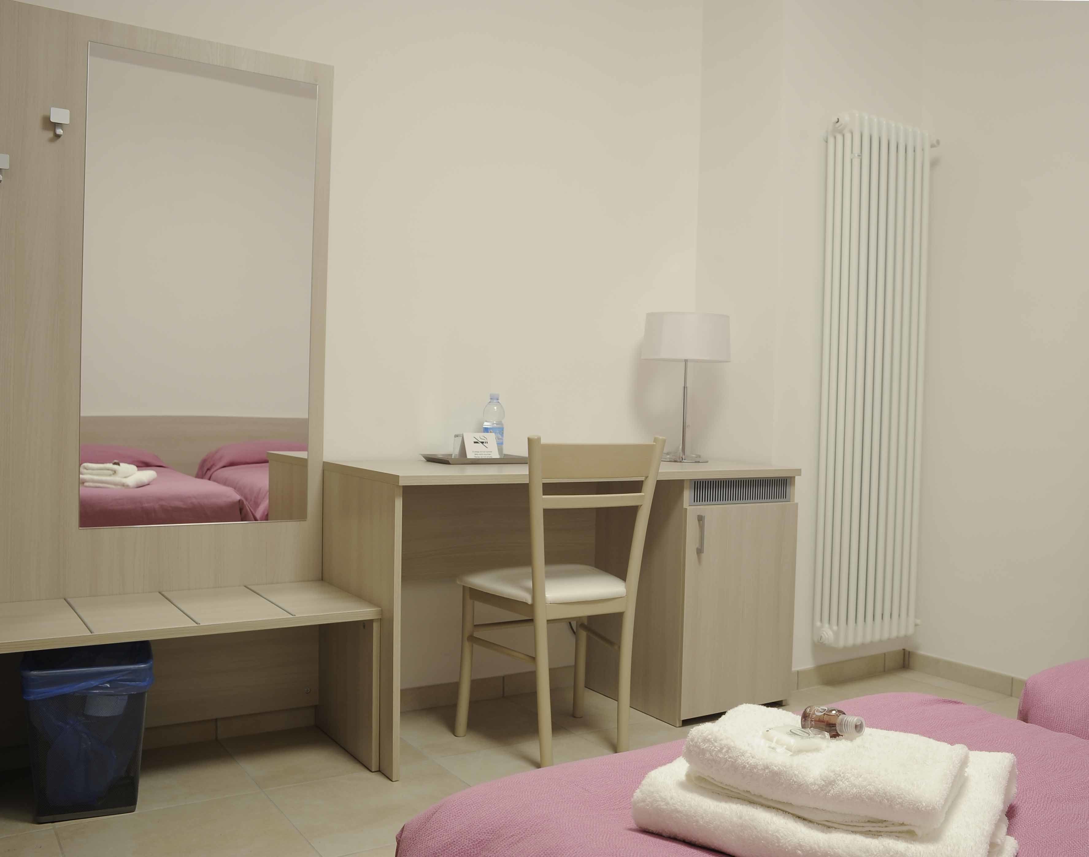 A hotel room with a bed, a desk with a chair, a mirror, a radiator, a lamp, and a trash bin.