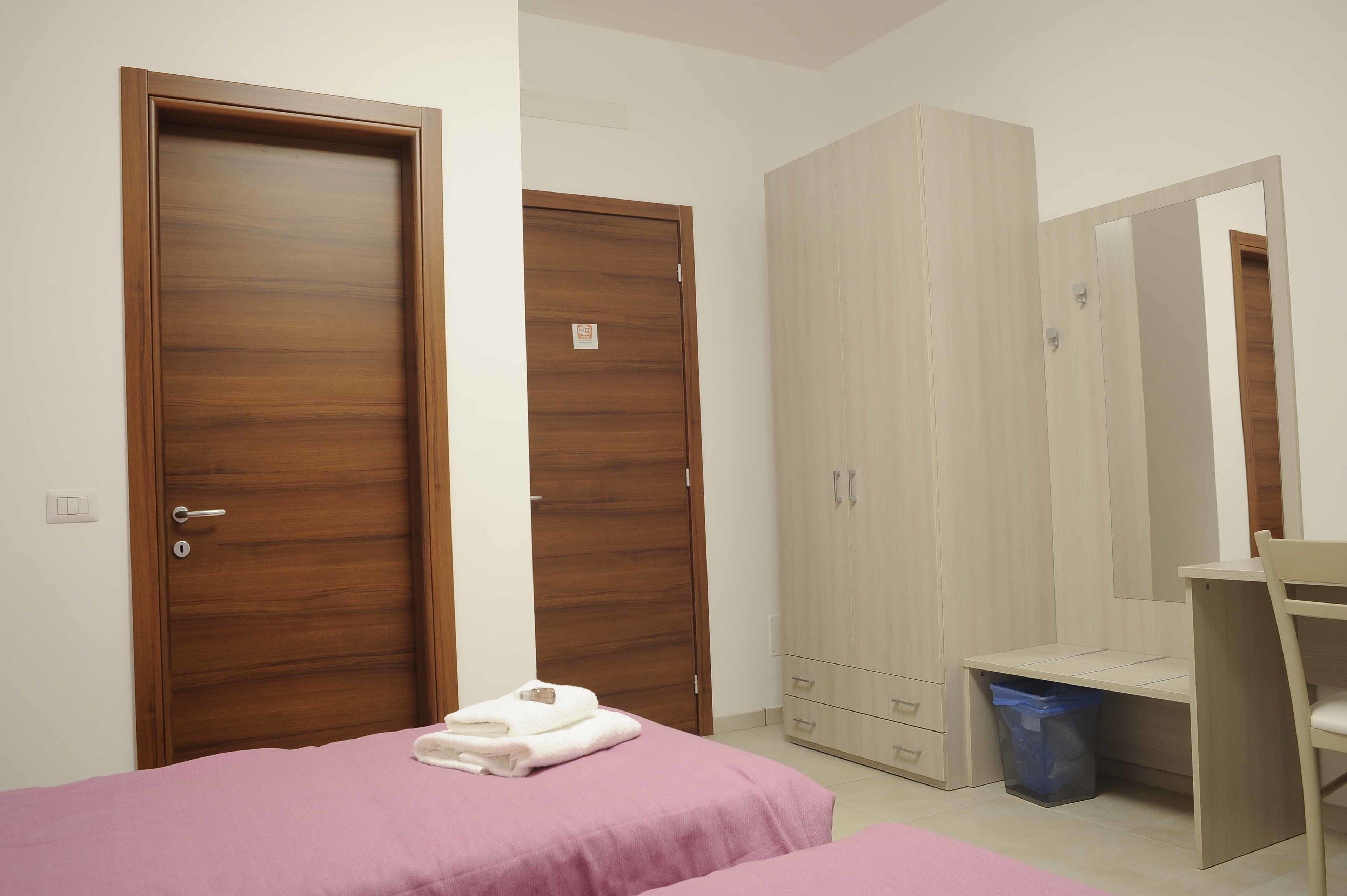 A bedroom with two wooden doors, a wardrobe, a desk, and a bed with pink bedding.