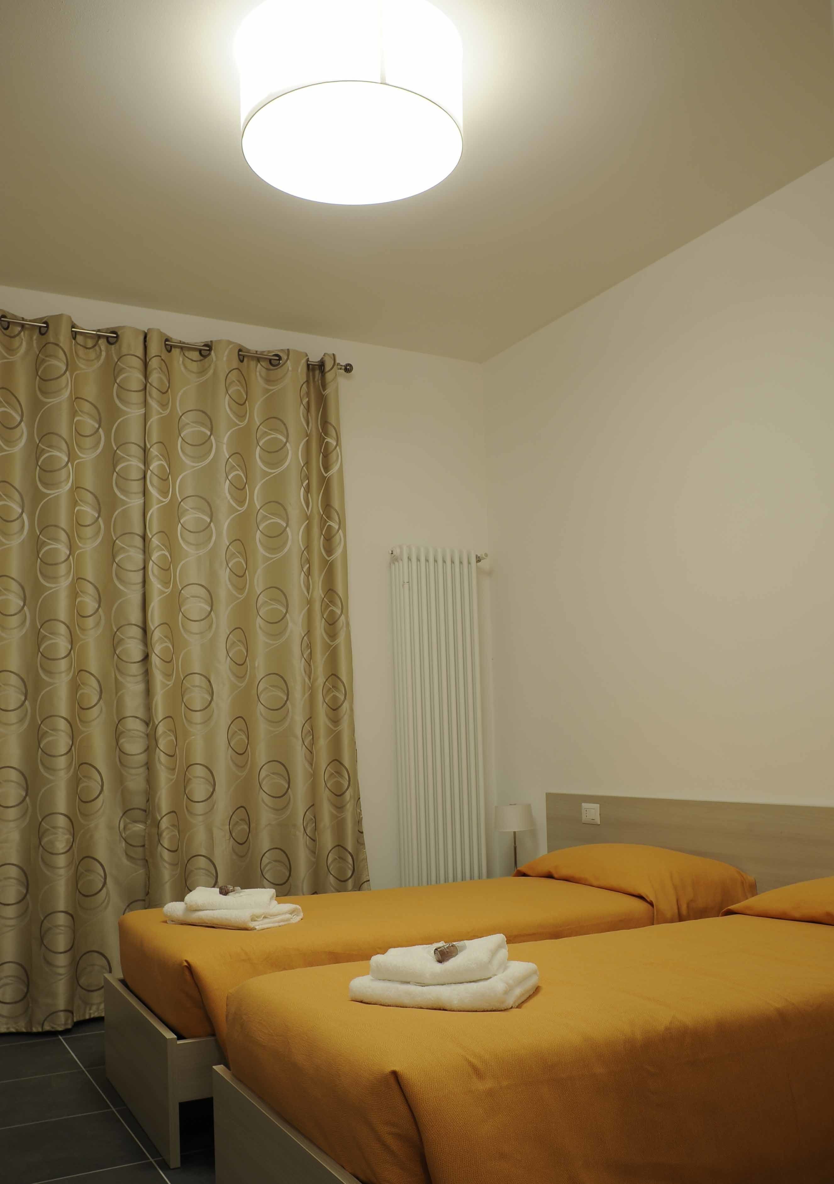 A hotel room with two beds, beige curtains, a white radiator, and a ceiling light.