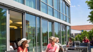 Un espace extérieur aménagé avec des tables et des chaises, un bâtiment moderne doté de grandes fenêtres et un ciel dégagé.