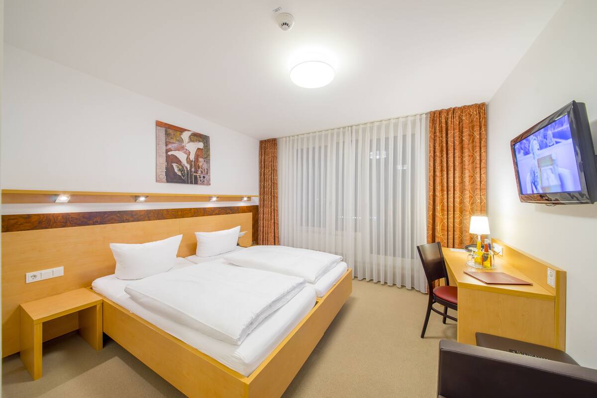 Ein Hotelzimmer mit Doppelbett, Schreibtisch mit Stuhl, Fernseher und einem Fenster mit Vorhängen.