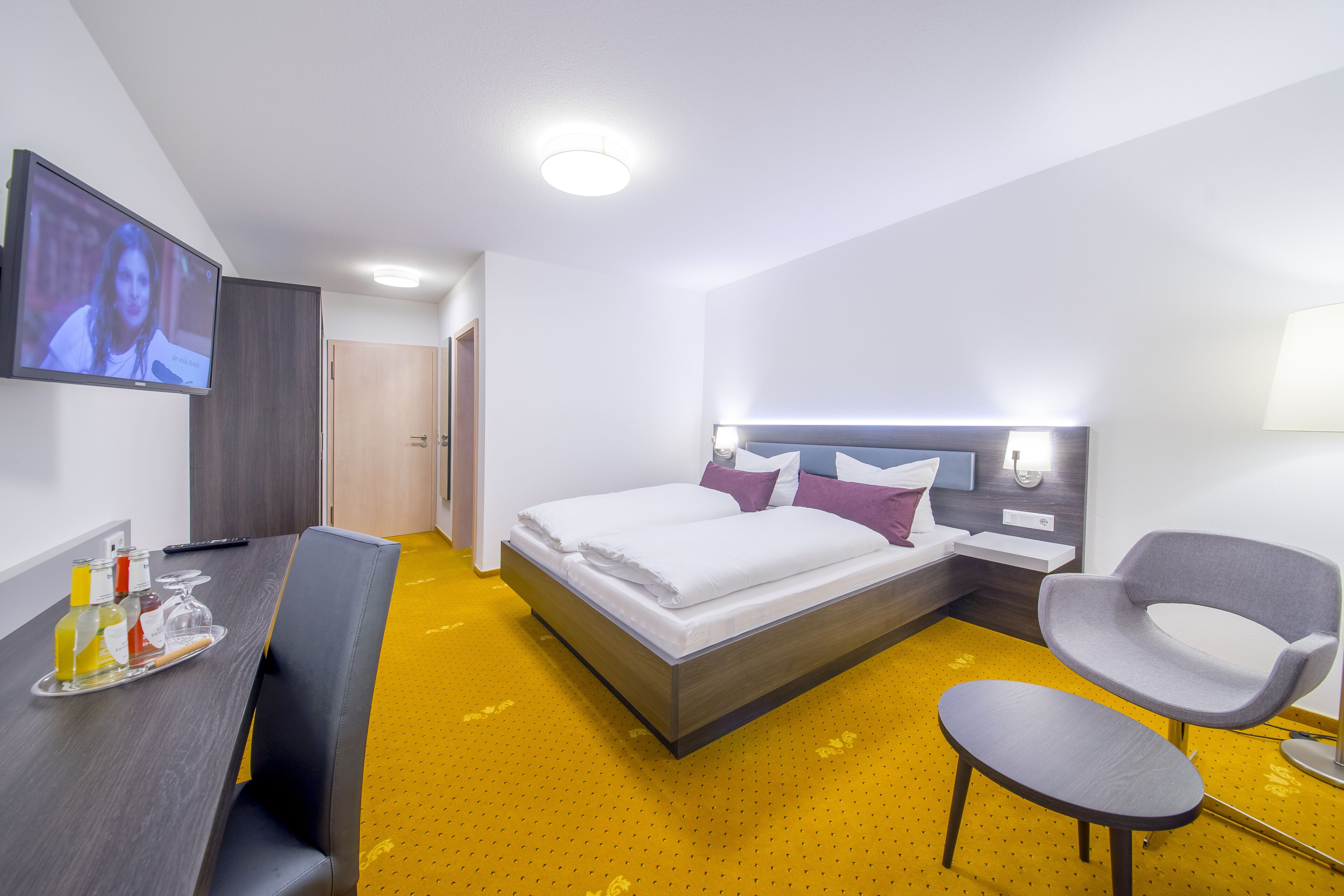 Una habitación de hotel con una cama, un escritorio con un neceser y una silla, y un televisor montado en la pared.