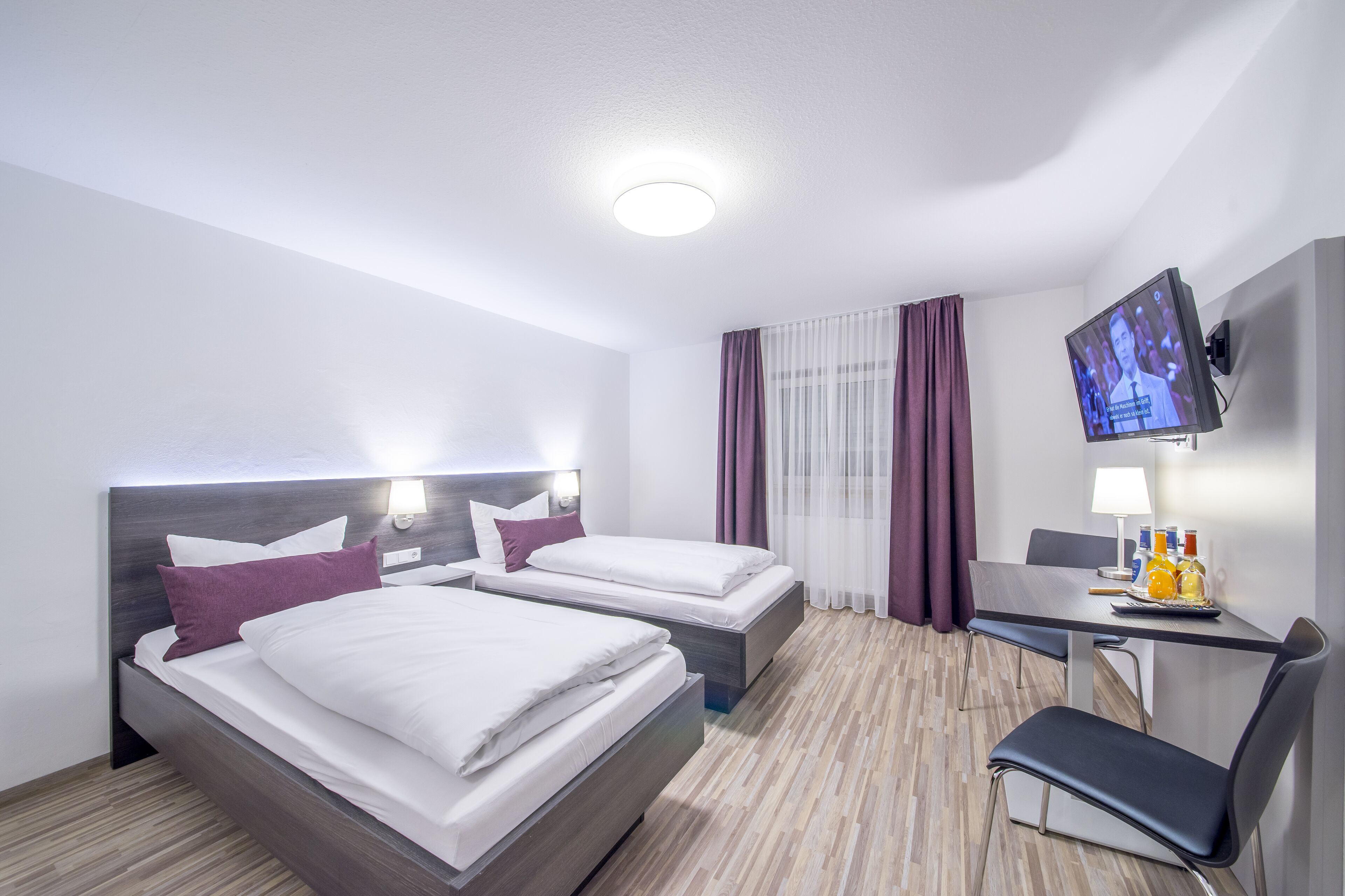 Una habitación de hotel con dos camas, un escritorio con silla, televisor y ventana con cortinas.