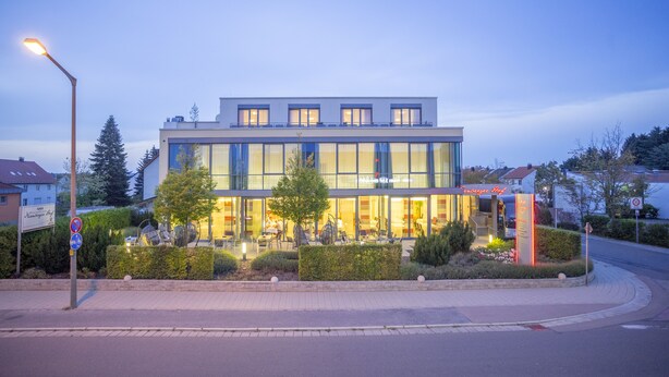 Un bâtiment moderne doté d’une façade en verre, d’un jardin aménagé et d’une enseigne indiquant la présence d’un restaurant.