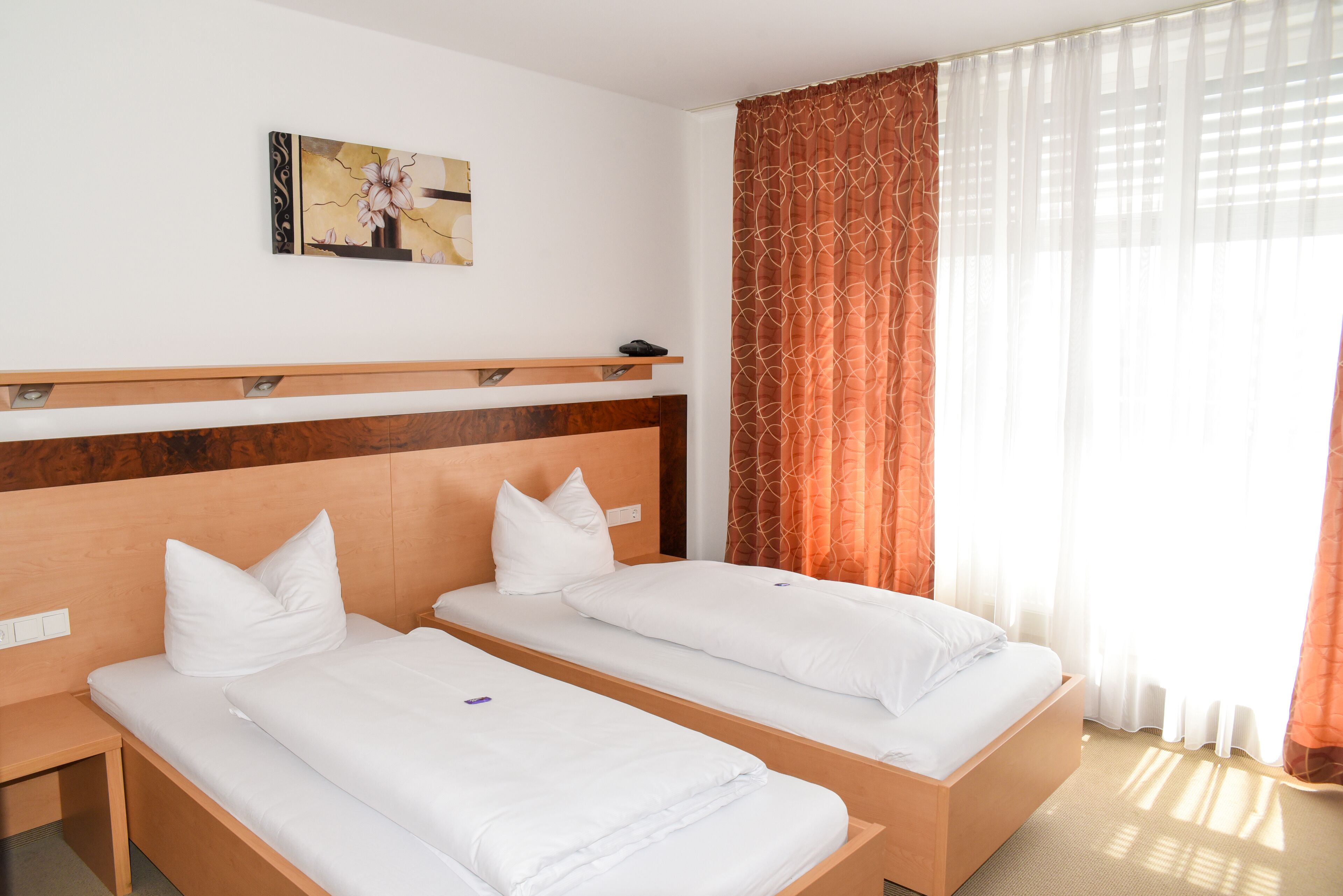 A hotel room with two single beds, a wooden headboard, a small table, a painting on the wall, and a window with sheer curtains.