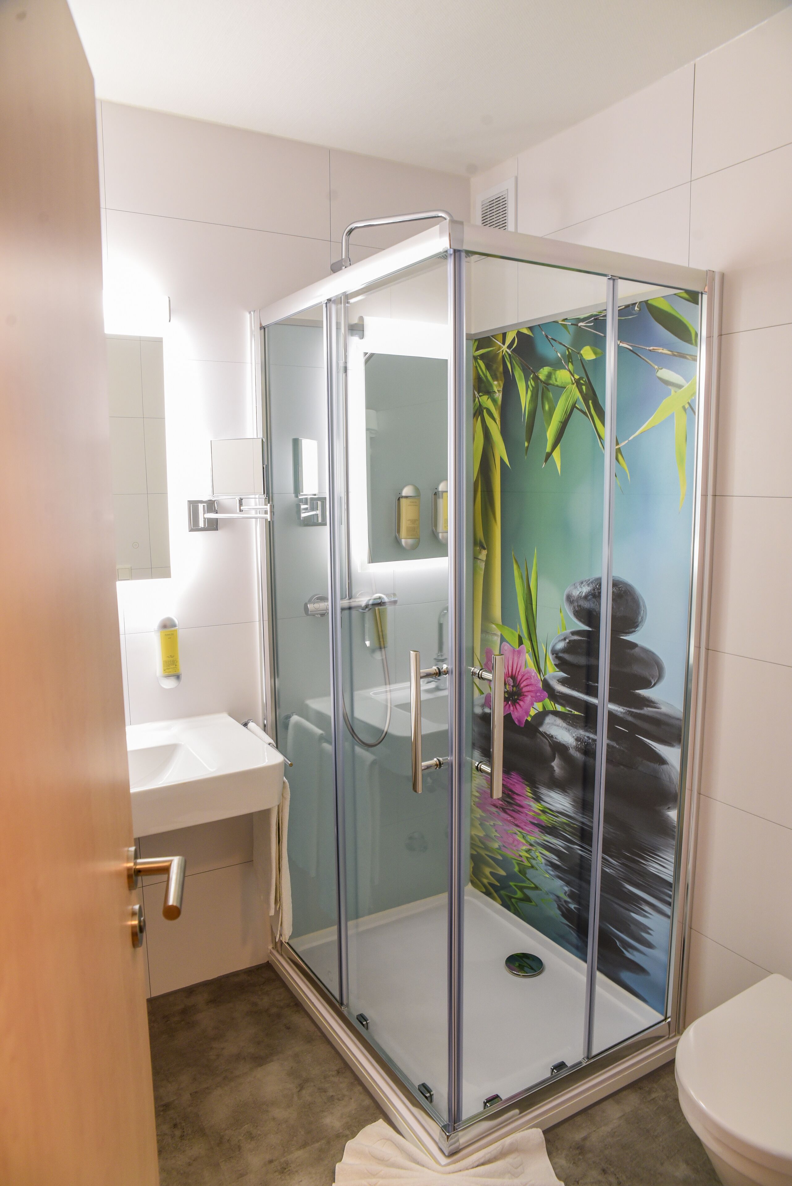 A glass-enclosed shower with a bamboo and water feature mural, a white sink, and a toilet.