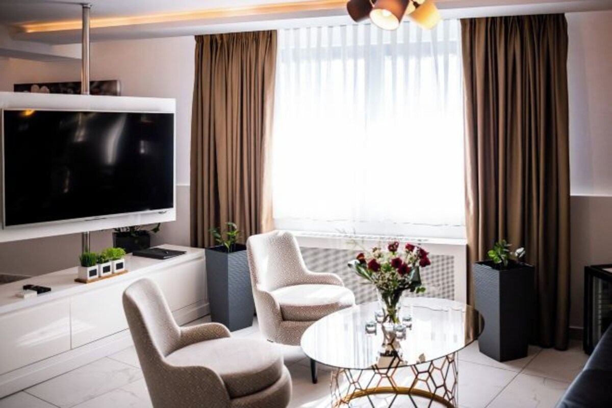 A modern living room with a flat-screen TV, two armchairs, a coffee table, and a large window with curtains.