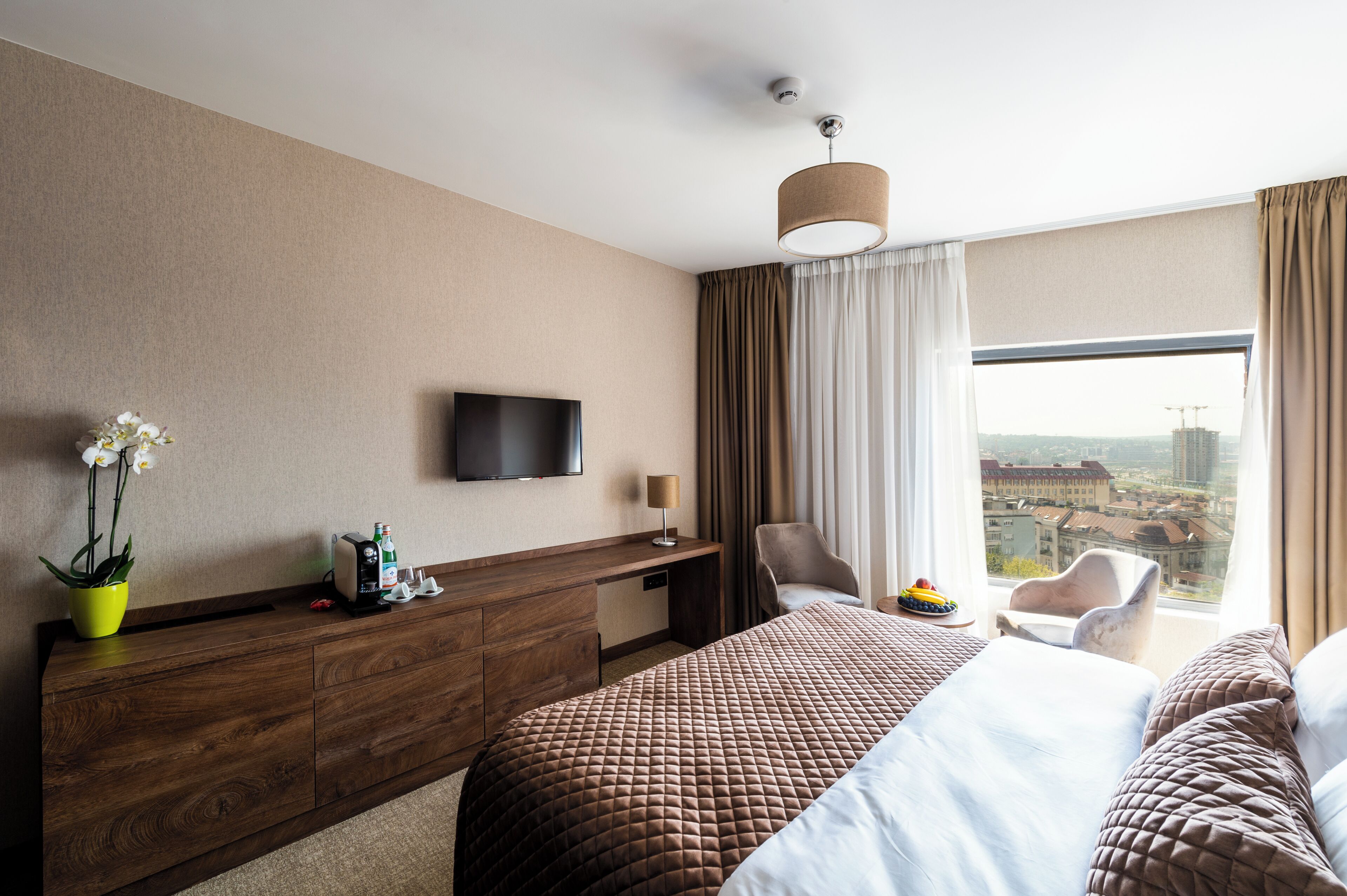 A hotel room with a large bed, a desk, a chair, a TV, and a window with a city view.