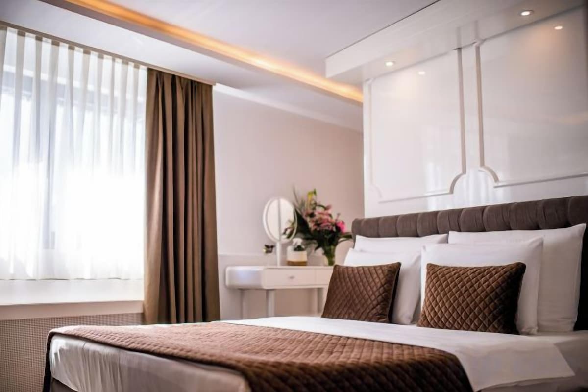 A modern hotel room with a large bed, a bedside table with a mirror, and a window with curtains.
