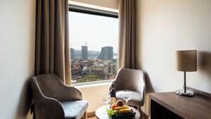 A hotel room with two chairs, a table with fruit and wine, and a view of a cityscape through a large window. - Amsterdam Hotel (Belgrade)