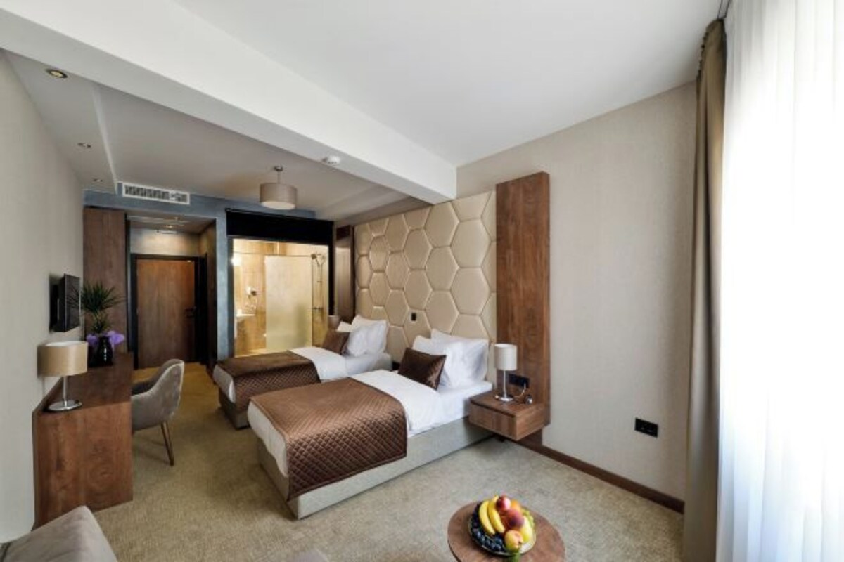 A hotel room with a large bed, two bedside tables, a desk with a chair, and a fruit bowl on the floor.