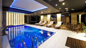 An indoor swimming pool with a blue-lit ceiling, lounge chairs, and a wooden table. - Amsterdam Hotel (Belgrade)