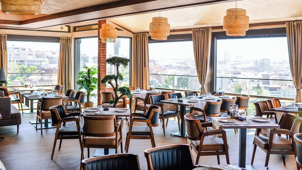 A spacious restaurant with high ceilings, large windows, and a city view. Tables are set with chairs, and there are hanging wicker lamps. - Amsterdam Hotel (Belgrade)