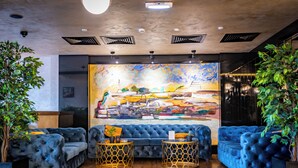 A modern lounge area with a large abstract painting, tufted blue sofas, and gold-patterned side tables. - Amsterdam Hotel (Belgrade)