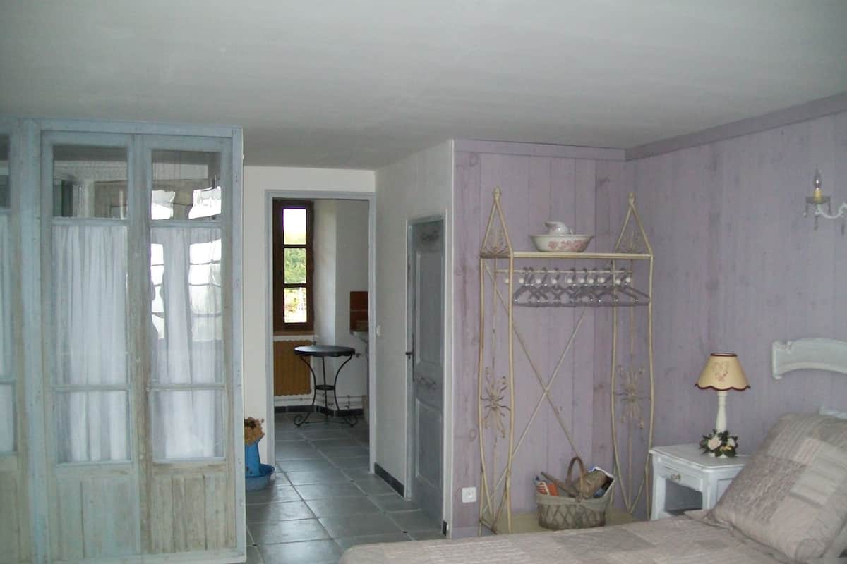 A bedroom with a bed, a nightstand, a lamp, a shelf with a bowl, and a chair in the background.