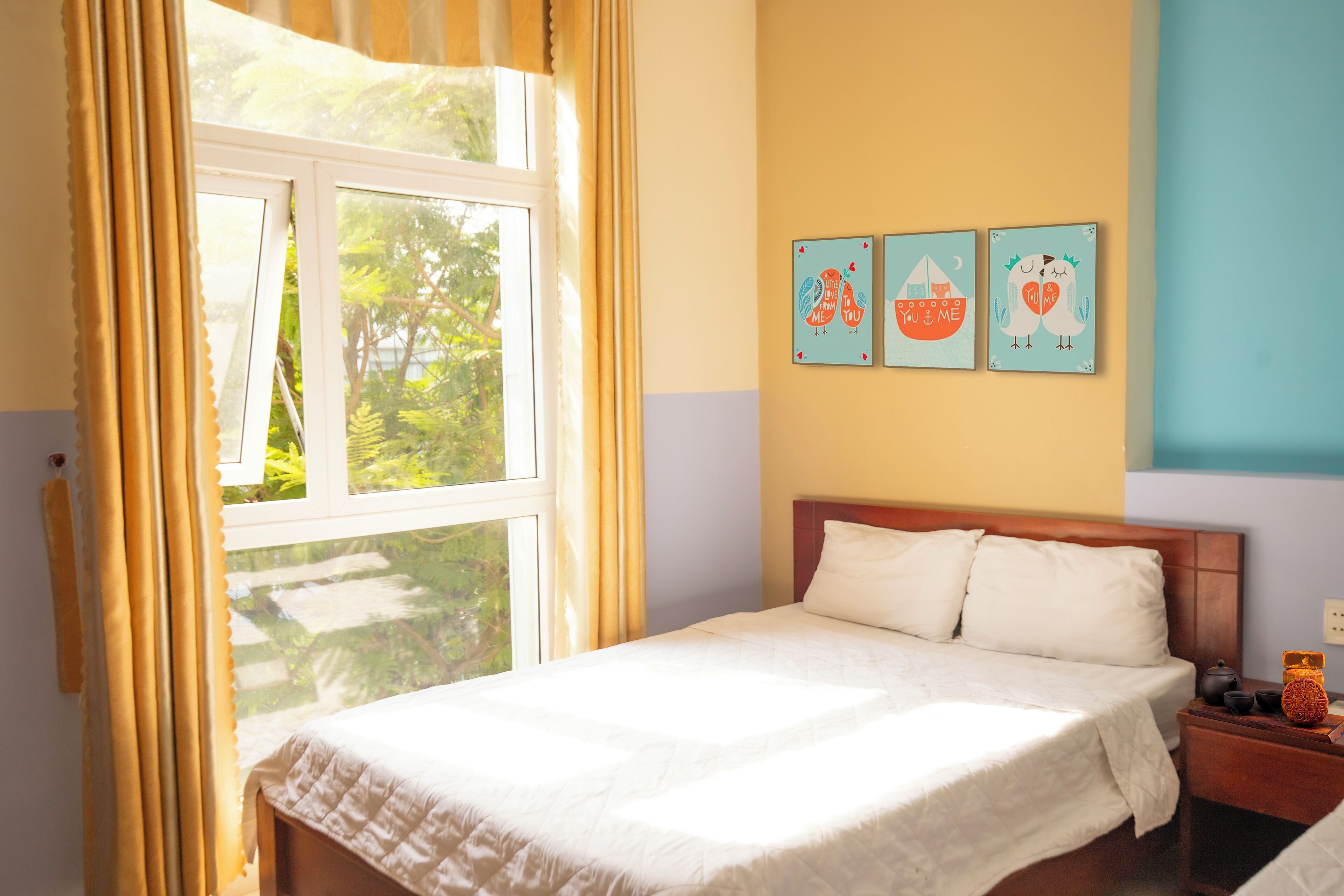 Un dormitorio con cama, mesita de noche y ventana con cortinas. Tres cuadros decoran la pared sobre la cama.