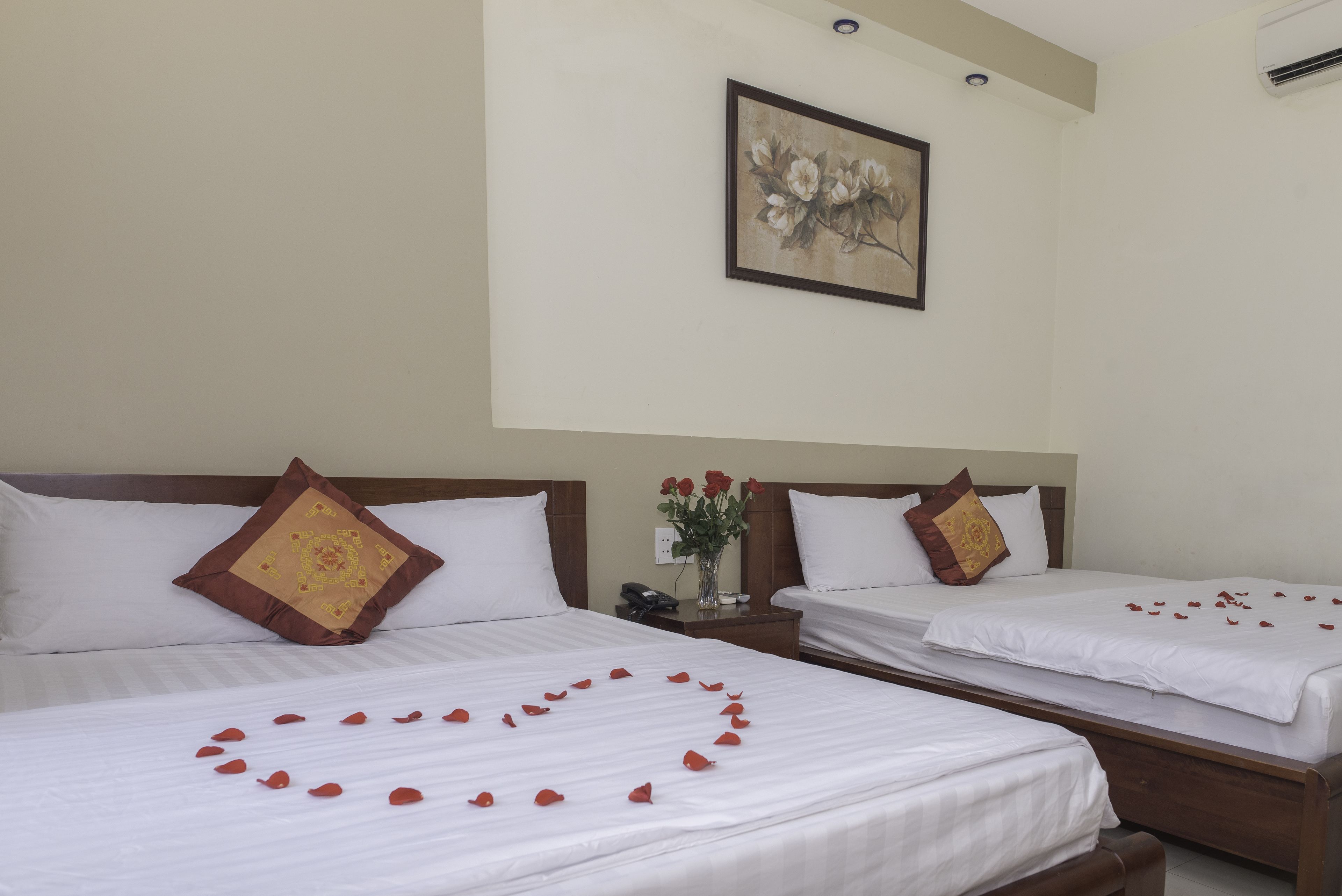 A hotel room with two beds, each with white bedding and a heart-shaped arrangement of rose petals on the bed in the foreground. There is a painting on the wall above the beds, and a vase with red roses on the nightstand between the beds.