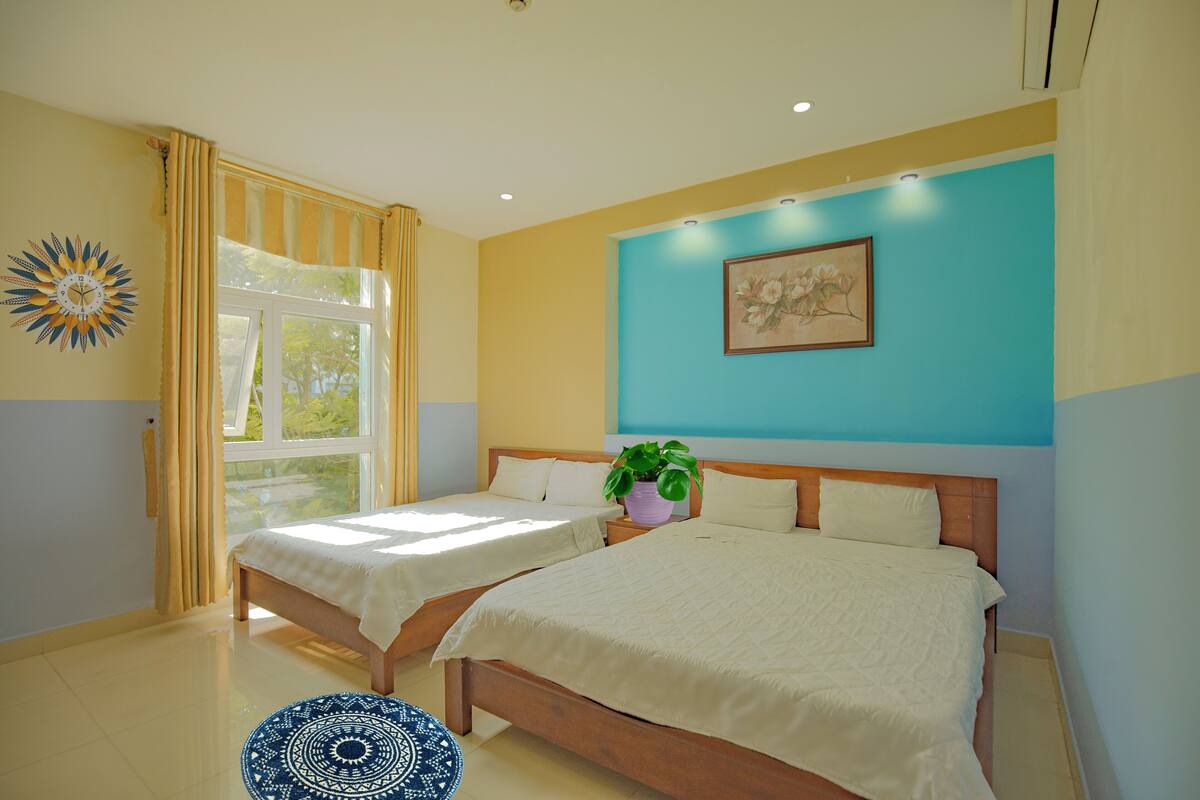 A hotel room with two beds, a large window with curtains, a potted plant, and a painting on the wall.