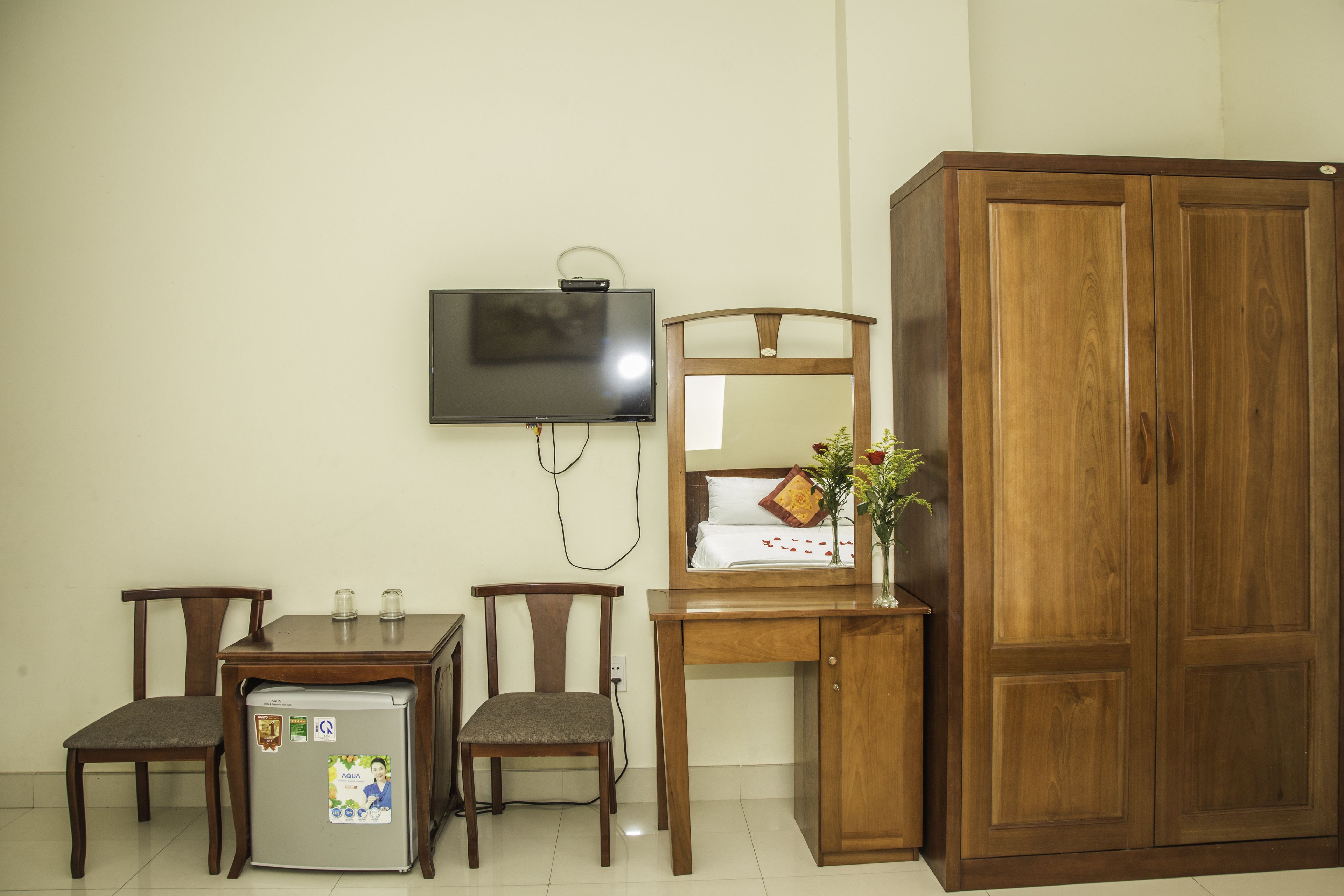 A room with a wooden wardrobe, a TV mounted on the wall, a small table with two chairs, and a refrigerator.