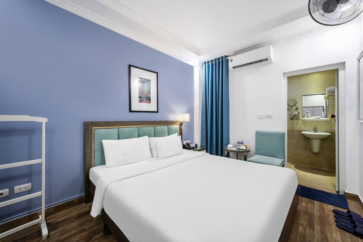 Habitación de hotel con cama, una silla, escritorio, equipo de aire acondicionado, lavamanos y espejo.