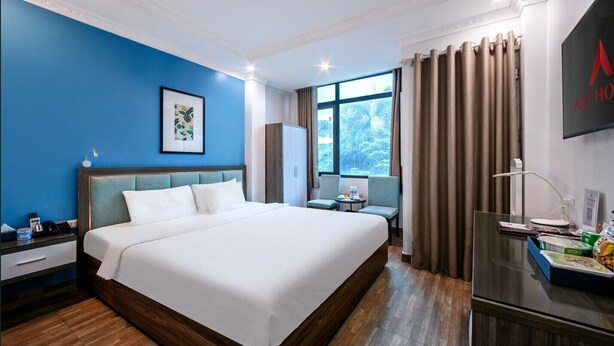 A hotel room with a large bed, a desk with a lamp, a chair, a window with curtains, and a TV on the wall. - A25 Hotel - 109 Truc Bach (Hanoi)