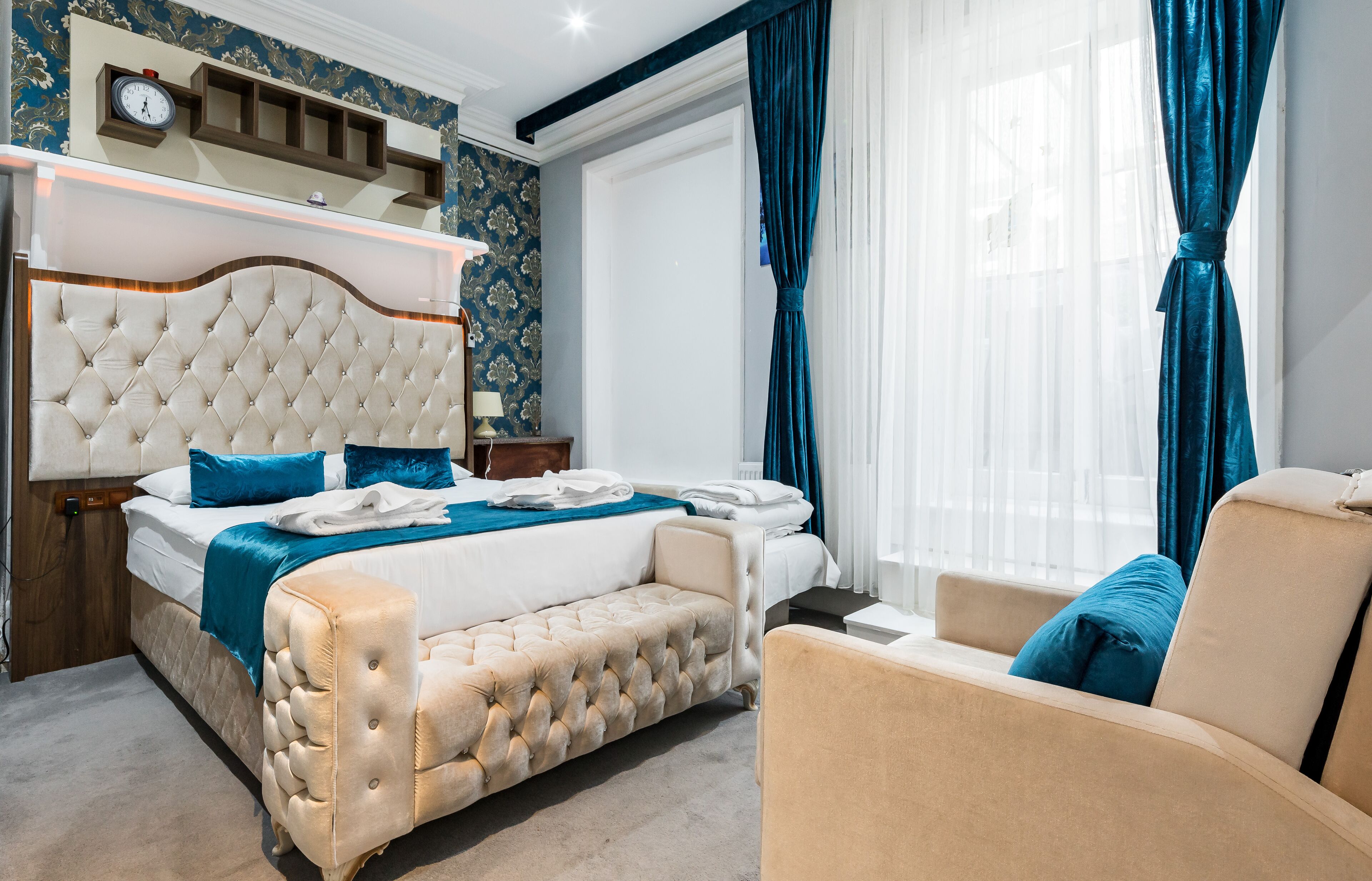 Un dormitorio con cabecera capitonada, cama con ropa de cama azul, mesita de noche con lámpara y ventana con cortinas.