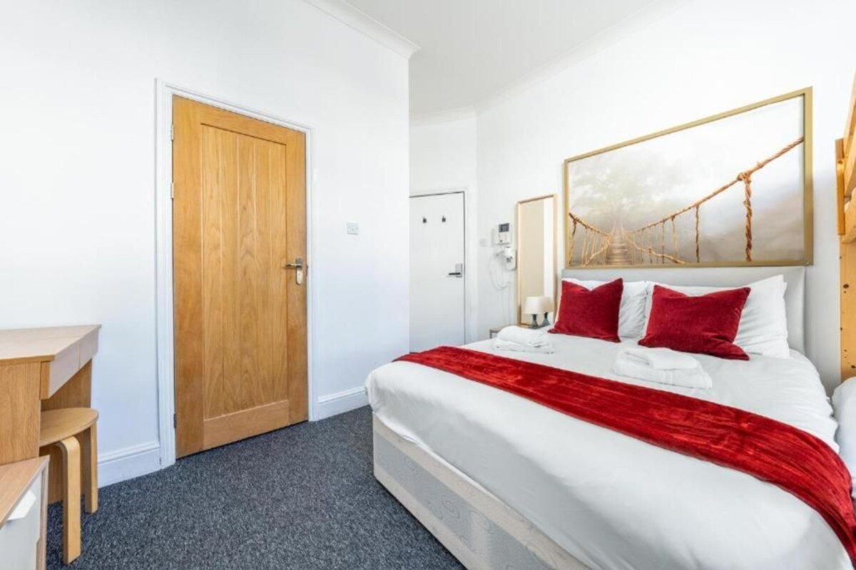 A hotel room with a bed, a desk, a wooden door, and a painting on the wall.