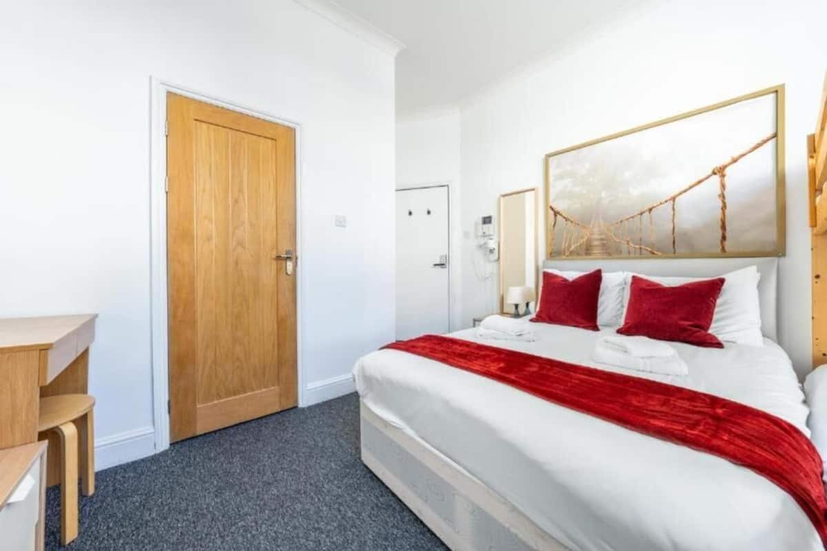 A hotel room with a bed, a desk, a wooden door, and a painting on the wall.
