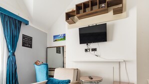 A hotel room with a beige armchair, a small round table, a TV mounted on the wall, and a wooden shelf above the TV. - Vilenza Hotel (London)