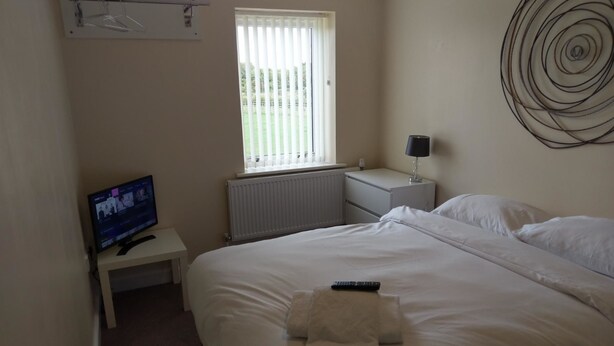 A hotel room with a bed, a TV on a small table, a window with blinds, a wall art piece, and a lamp on a bedside table. - Jaylets Budget Glenfield (Leicester)