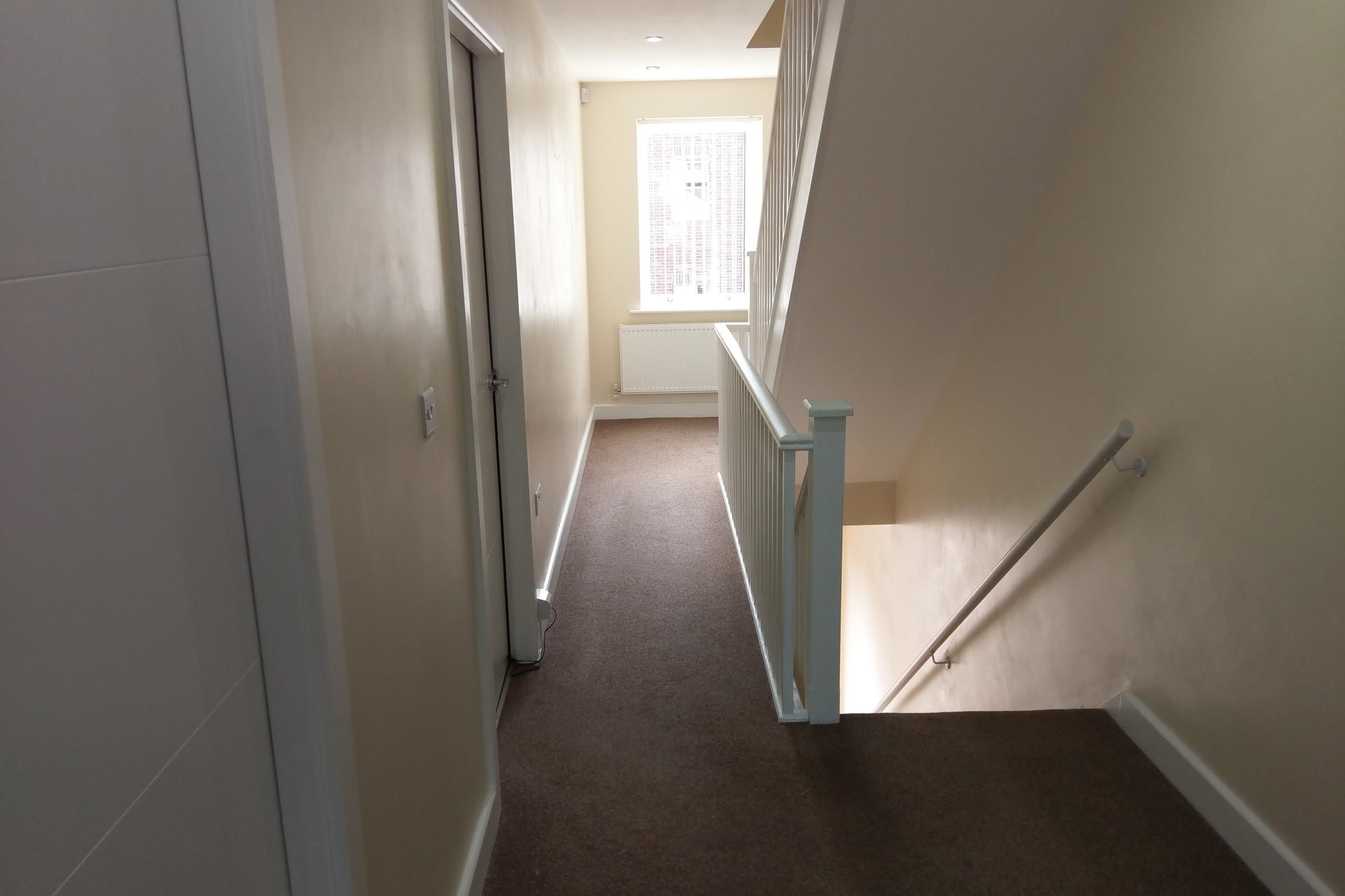 A narrow, carpeted hallway with a white door on the left, a white railing on the right, and a window at the end.