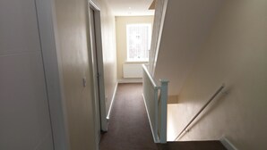 A narrow, carpeted hallway with a white door on the left, a white railing on the right, and a window at the end. - Jaylets Budget Glenfield (Leicester)