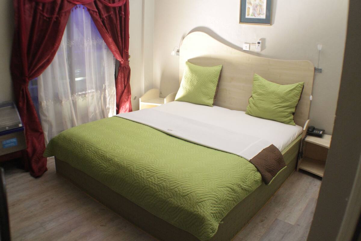 A neatly made bed with green and white bedding, a bedside table with a lamp, and a red curtain partially drawn.