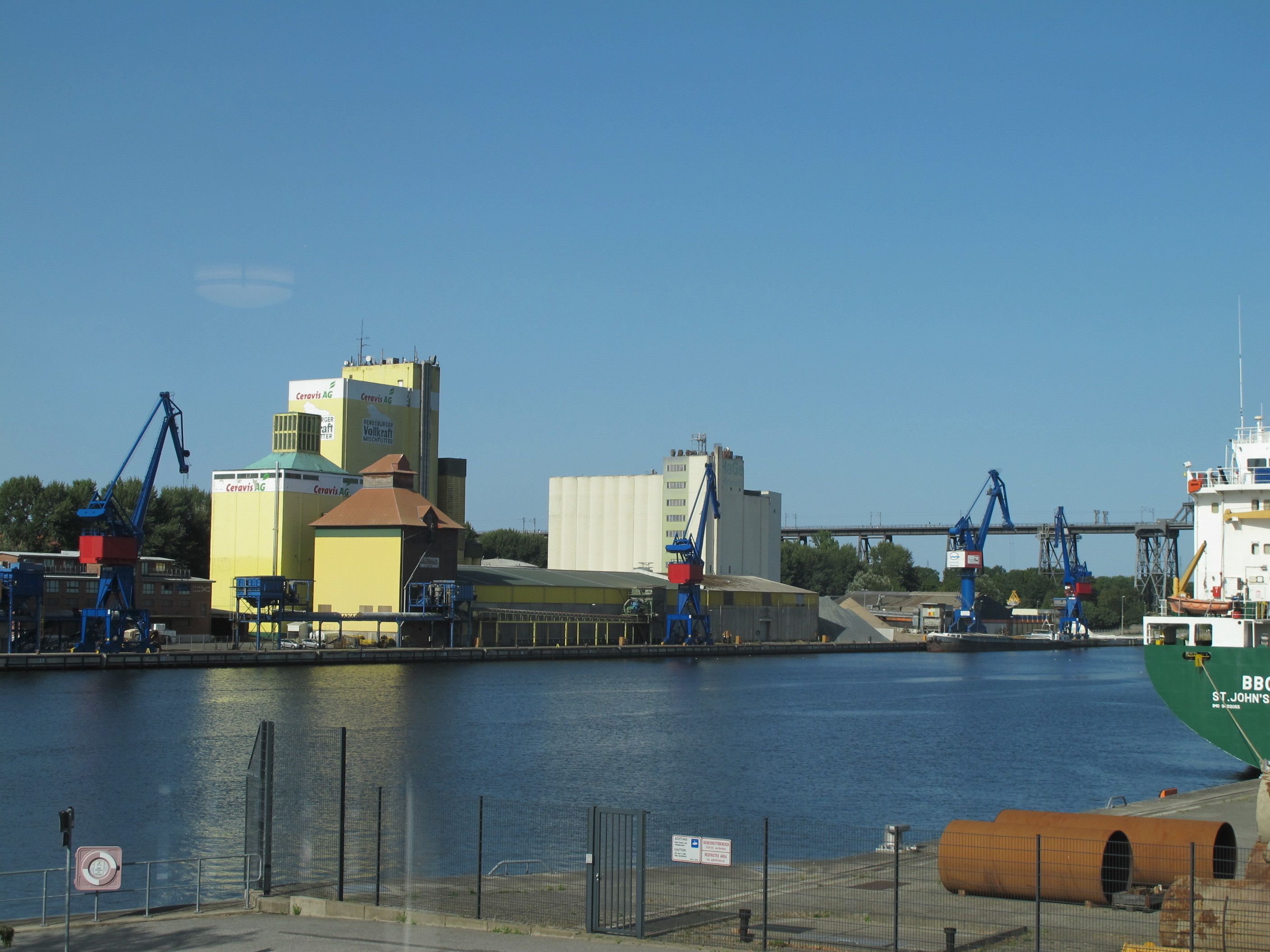 A waterfront industrial area with cranes, a large green ship, and buildings with signage.