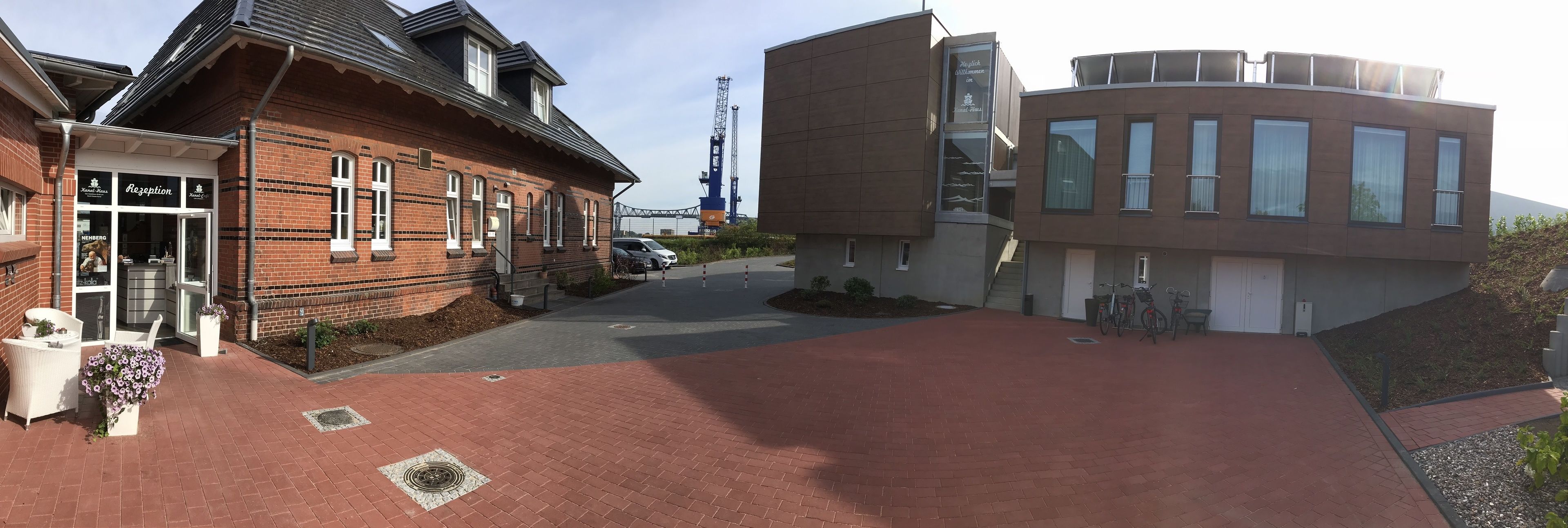 A reception area with a brick building, a modern structure with large windows, and a pathway leading to a parking area.