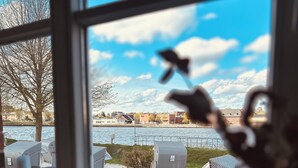 A view through a window of outdoor wicker furniture and chairs by a body of water, with buildings in the background. - Kanal-Haus "Das Gästehaus direkt am Nord-Ostsee-Kanal" (Osterrönfeld)