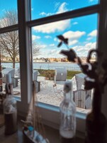 Blick durch ein Fenster auf Gartenmöbel aus Rattan und Stühle an einem Gewässer, mit Gebäuden im Hintergrund.