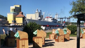 Beach huts with striped awnings, a large ship, and industrial buildings in the background. - Kanal-Haus "Das Gästehaus direkt am Nord-Ostsee-Kanal" (Osterrönfeld)