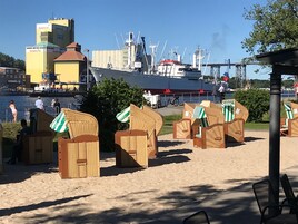 Strandhütten mit Streifenmarkisen, ein großes Schiff und im Hintergrund Industriegebäude.