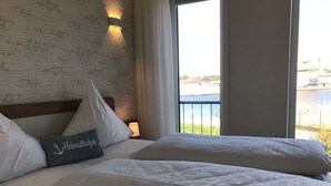 A hotel room with two beds, a large window with a view of a body of water and buildings, and a wall with signatures. - Kanal-Haus "Das Gästehaus direkt am Nord-Ostsee-Kanal" (Osterrönfeld)