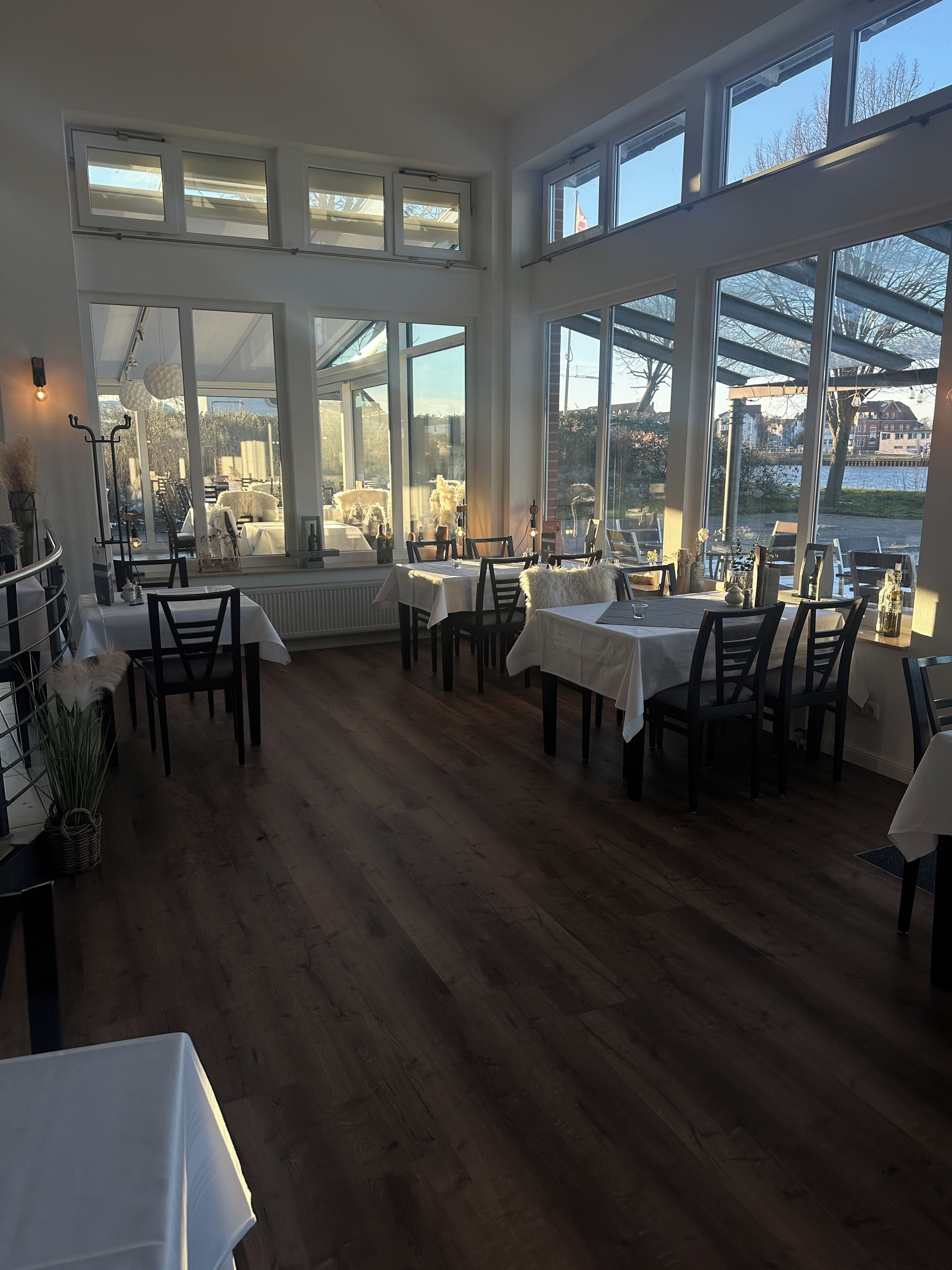 A dining area with multiple tables and chairs, large windows with views of a body of water and buildings, and a wooden floor.