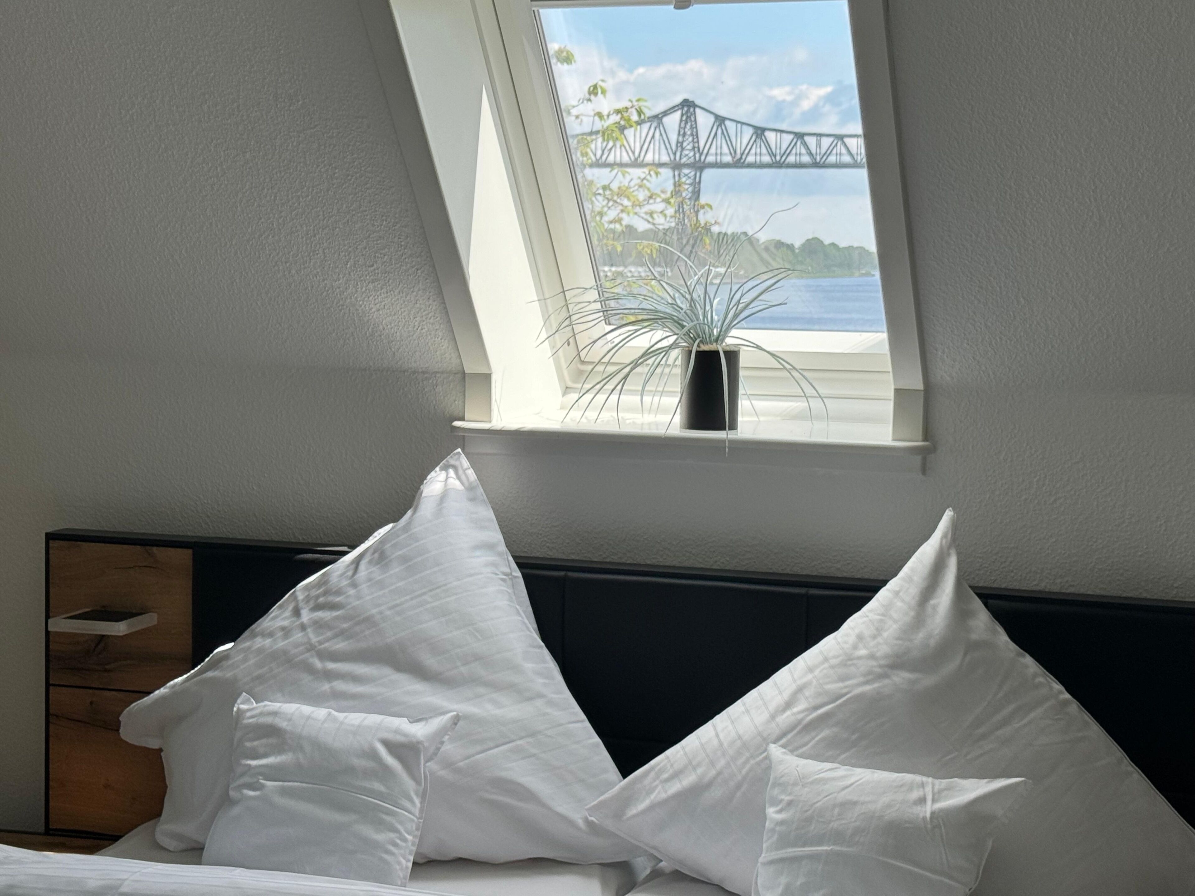 Une chambre à coucher avec un lit, des oreillers blancs, une table de chevet, une plante en pot et une fenêtre donnant sur un pont et sur l’eau.