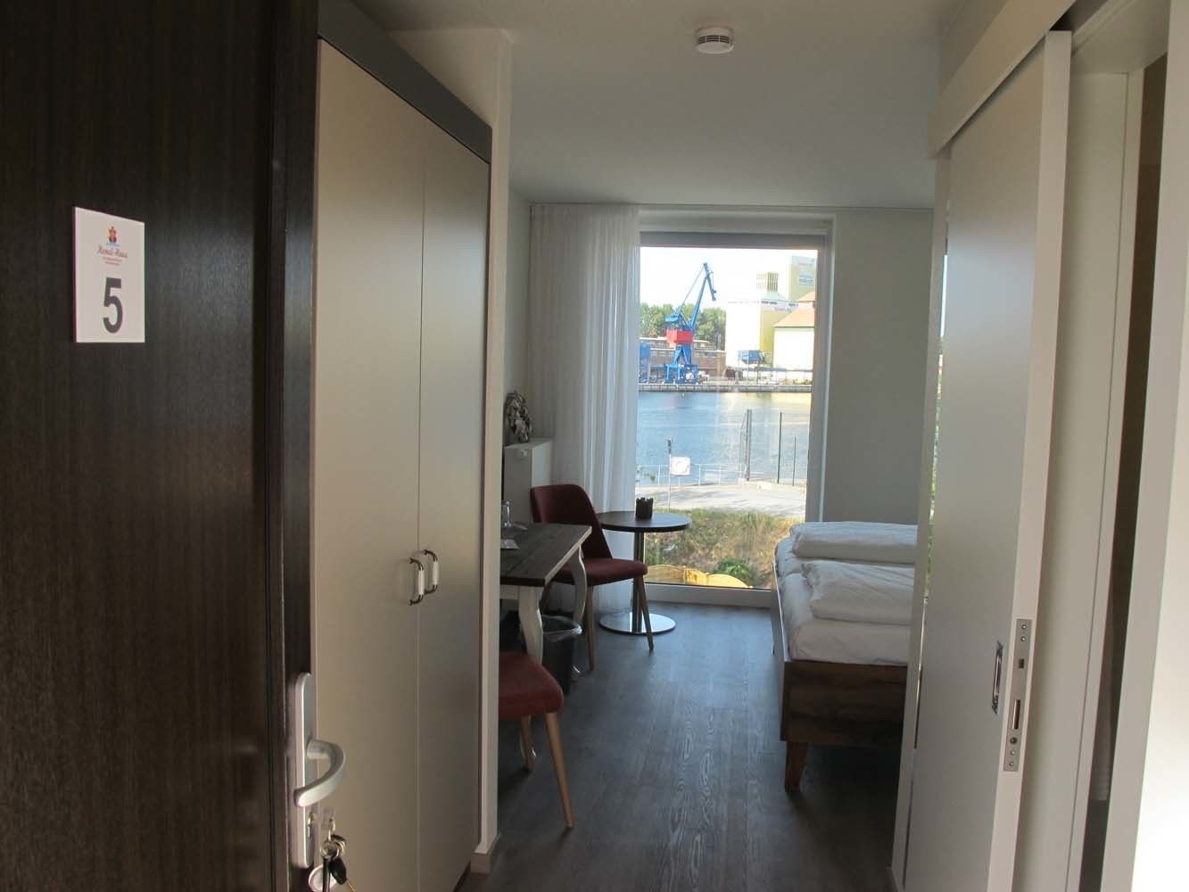 Una camera d'albergo con un letto, una piccola scrivania con una sedia, una finestra che dà su panorama esterno e una porta con il simbolo del numero.