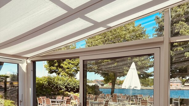 An outdoor dining area with tables and chairs, a view of a pool and umbrellas, and a covered section with a white ceiling. - Kanal-Haus "Das Gästehaus direkt am Nord-Ostsee-Kanal" (Osterrönfeld)