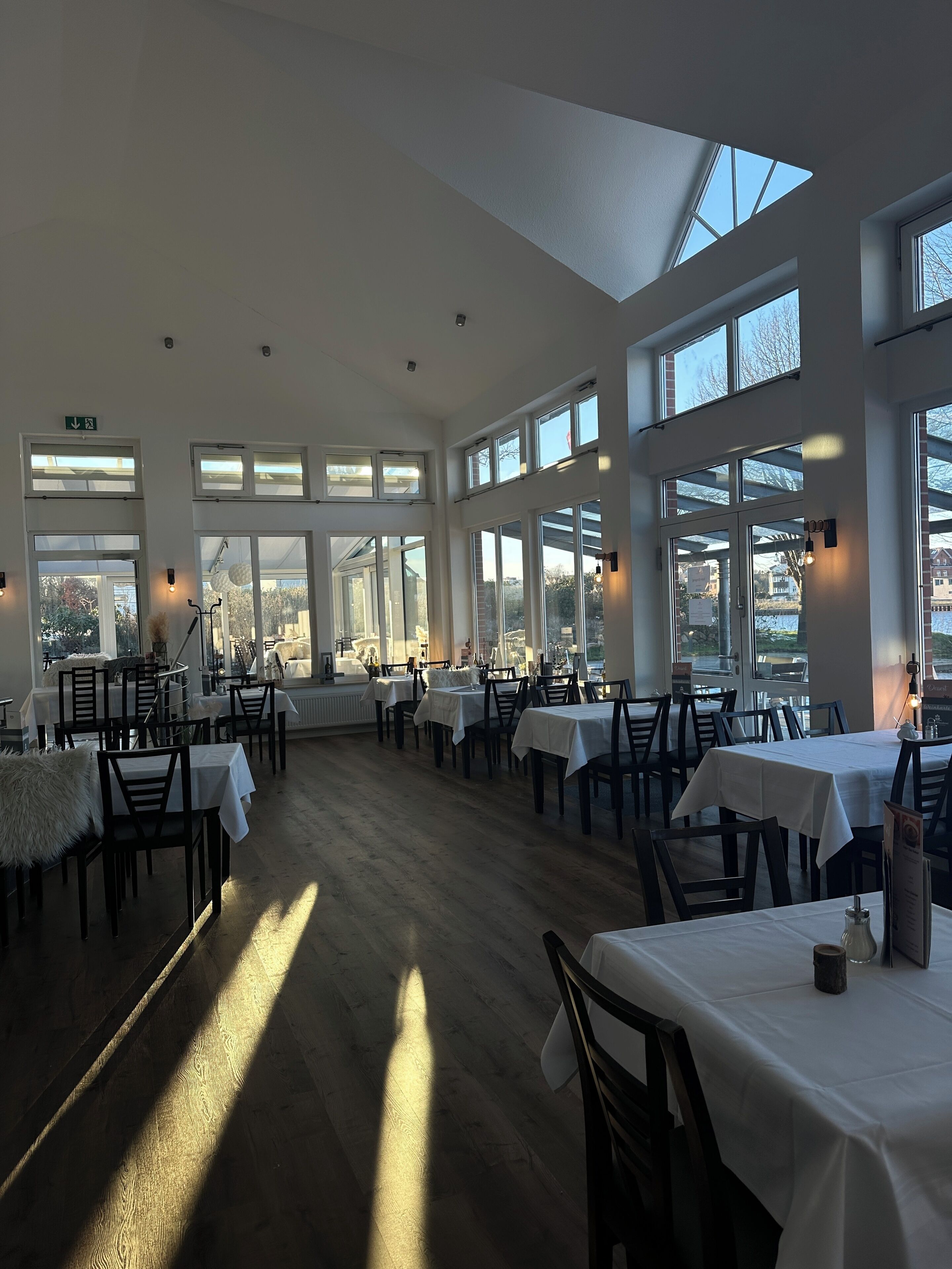 A spacious restaurant with high ceilings and large windows, allowing natural light to fill the room. Tables are neatly arranged, and the floor is wooden.